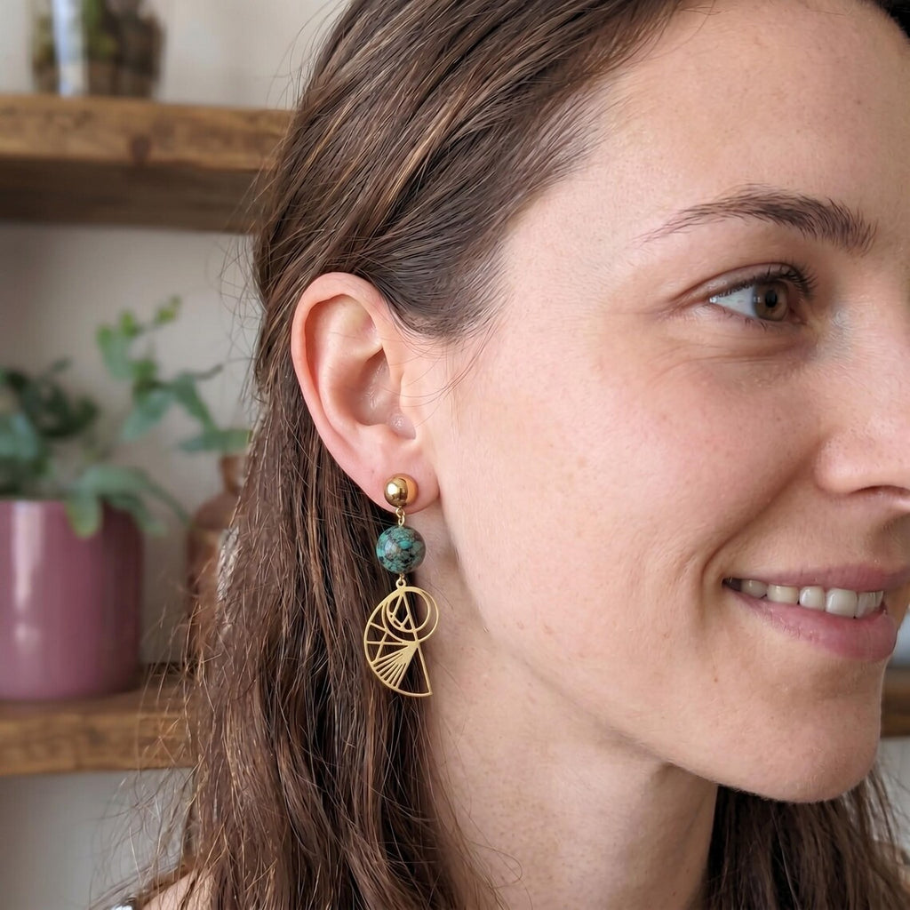 A close-up of a woman wearing Bohemian gold-plated stainless steel drop earrings featuring round, green-speckled African Turquoise beads and geometric Golden Ratio shell charms, set against a softly blurred indoor background.