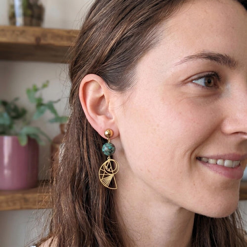 A close-up of a woman wearing Bohemian gold-plated stainless steel drop earrings featuring round, green-speckled African Turquoise beads and geometric Golden Ratio shell charms, set against a softly blurred indoor background.
