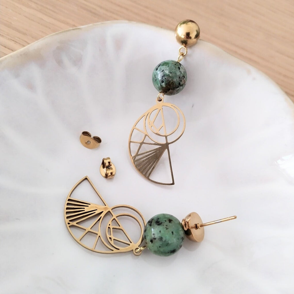 Bohemian gold-plated stainless steel drop earrings featuring round, green-speckled African Turquoise beads and geometric Golden Ratio shell charms, displayed on a white ceramic dish over a light wood surface.