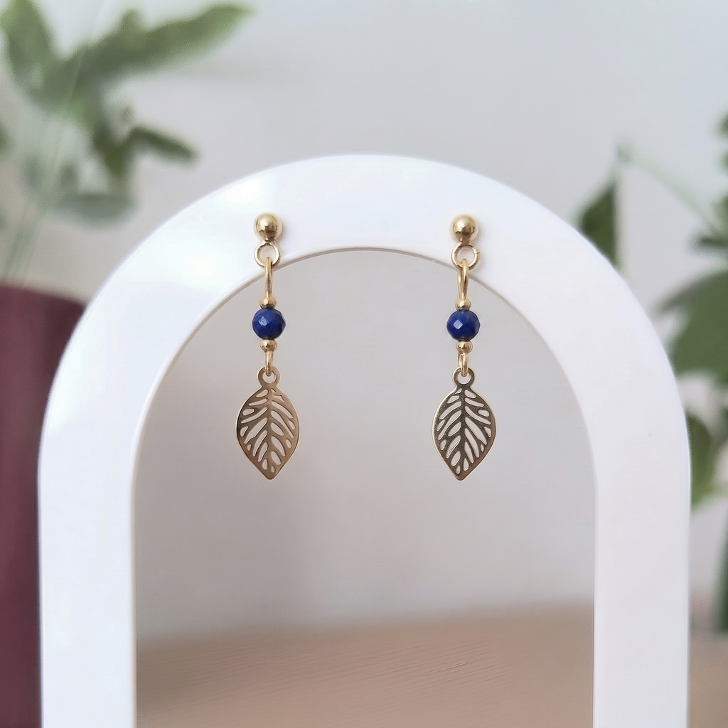 A pair of gold-toned drop earrings with a small round blue lapis lazuli bead and a leaf-shaped charm, displayed on a white arched stand with a softly blurred indoor background.