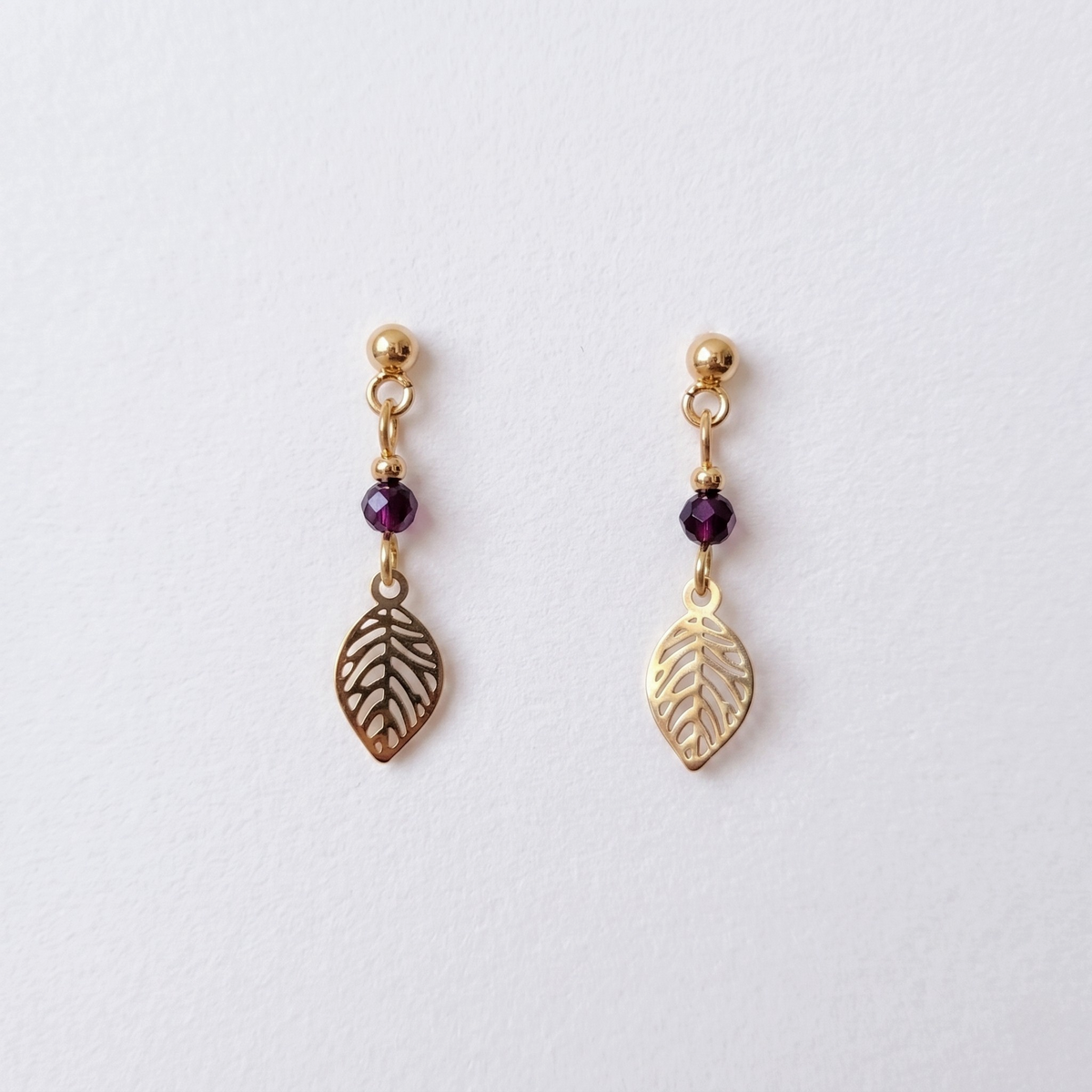 A pair of gold-toned dangle earrings with small faceted garnet beads and leaf-shaped charms, displayed on a clean white background.