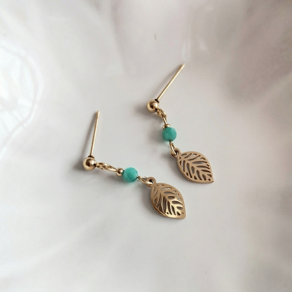 A pair of gold dangle earrings with leaf-shaped charms and small aqua amazonite beads, displayed on a soft white surface.