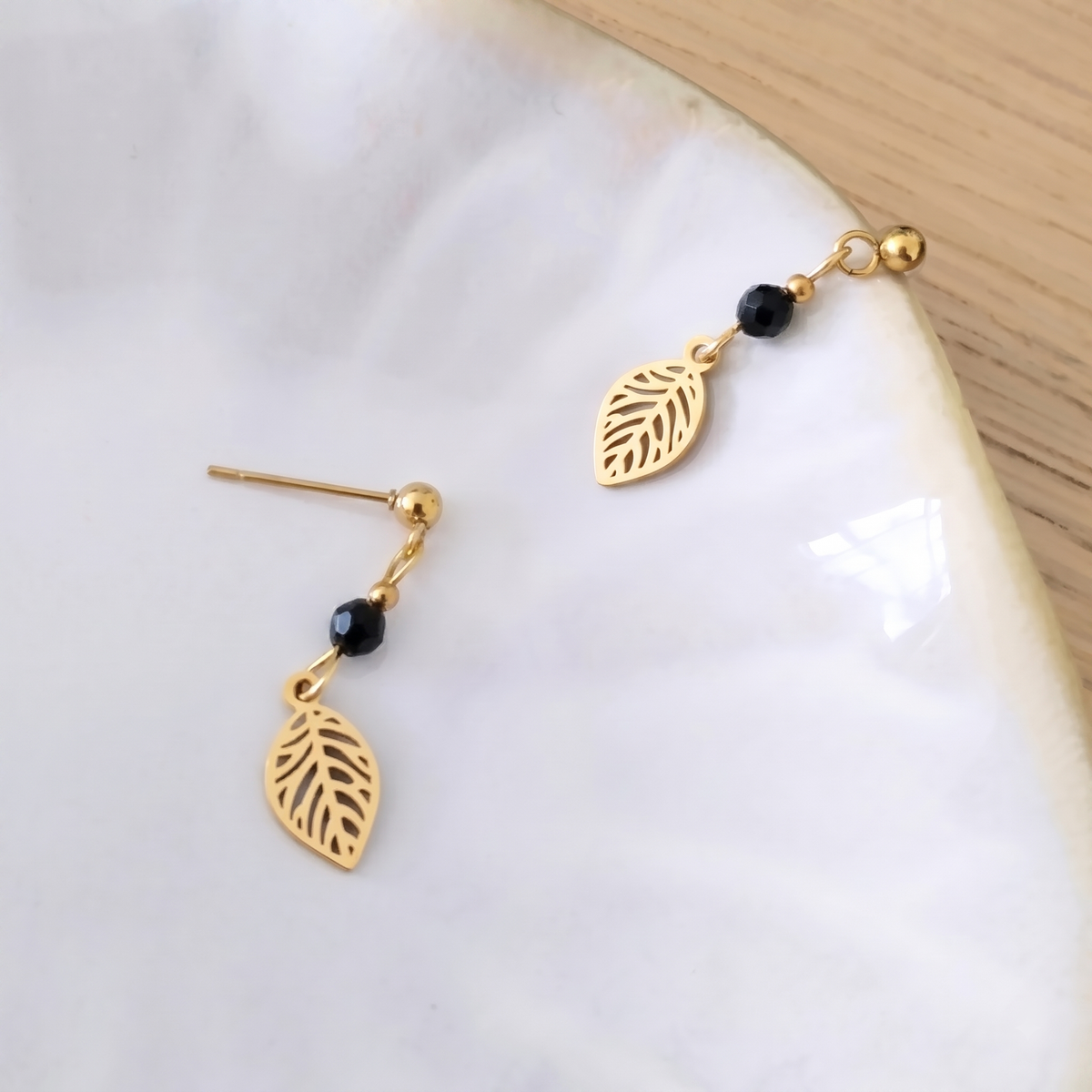 A pair of gold leaf drop earrings with small black obsidian beads, displayed on a white marble surface with a light wood background.
