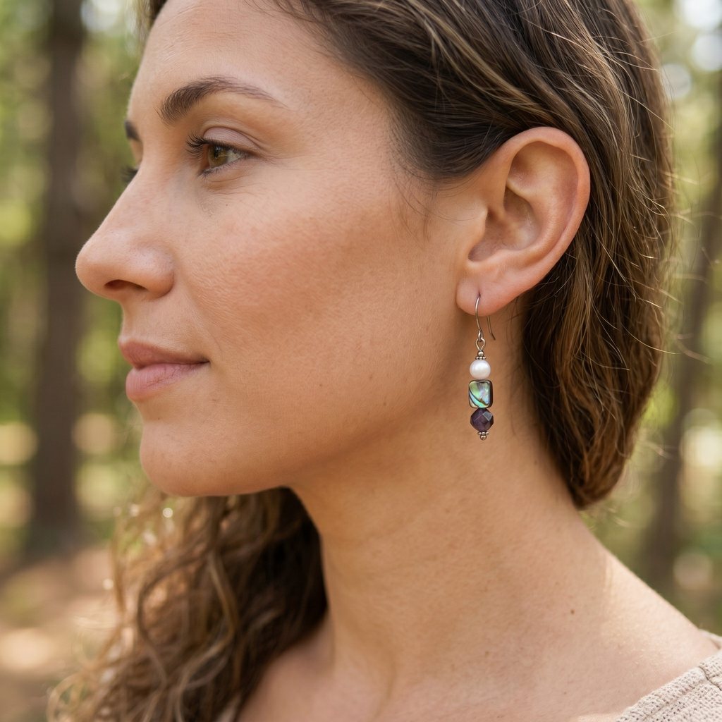 A close-up side profile of a woman wearing a dangling earring with a white pearl, a Paua bead, and an amethyst purple bead, set against a blurred forest background.